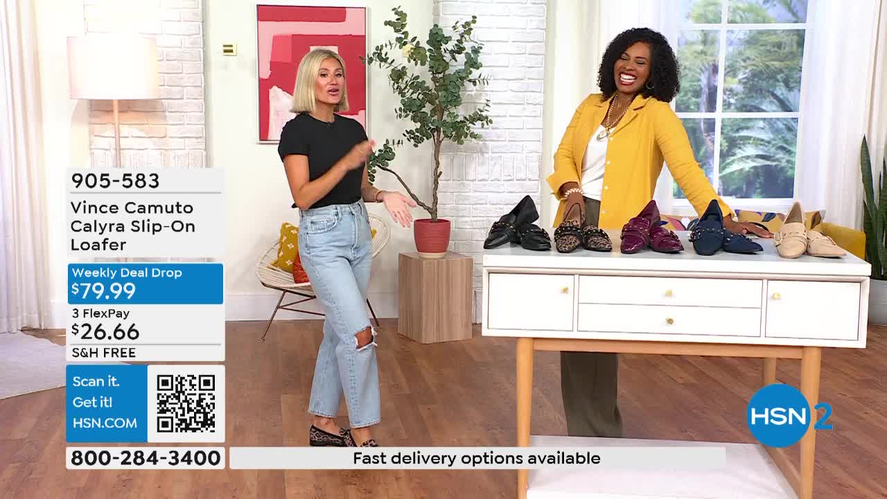Two women stand in a brightly lit studio, gesturing towards a selection of loafers displayed on a white table. The HSN2 logo is visible, and the screen displays product details and pricing.
