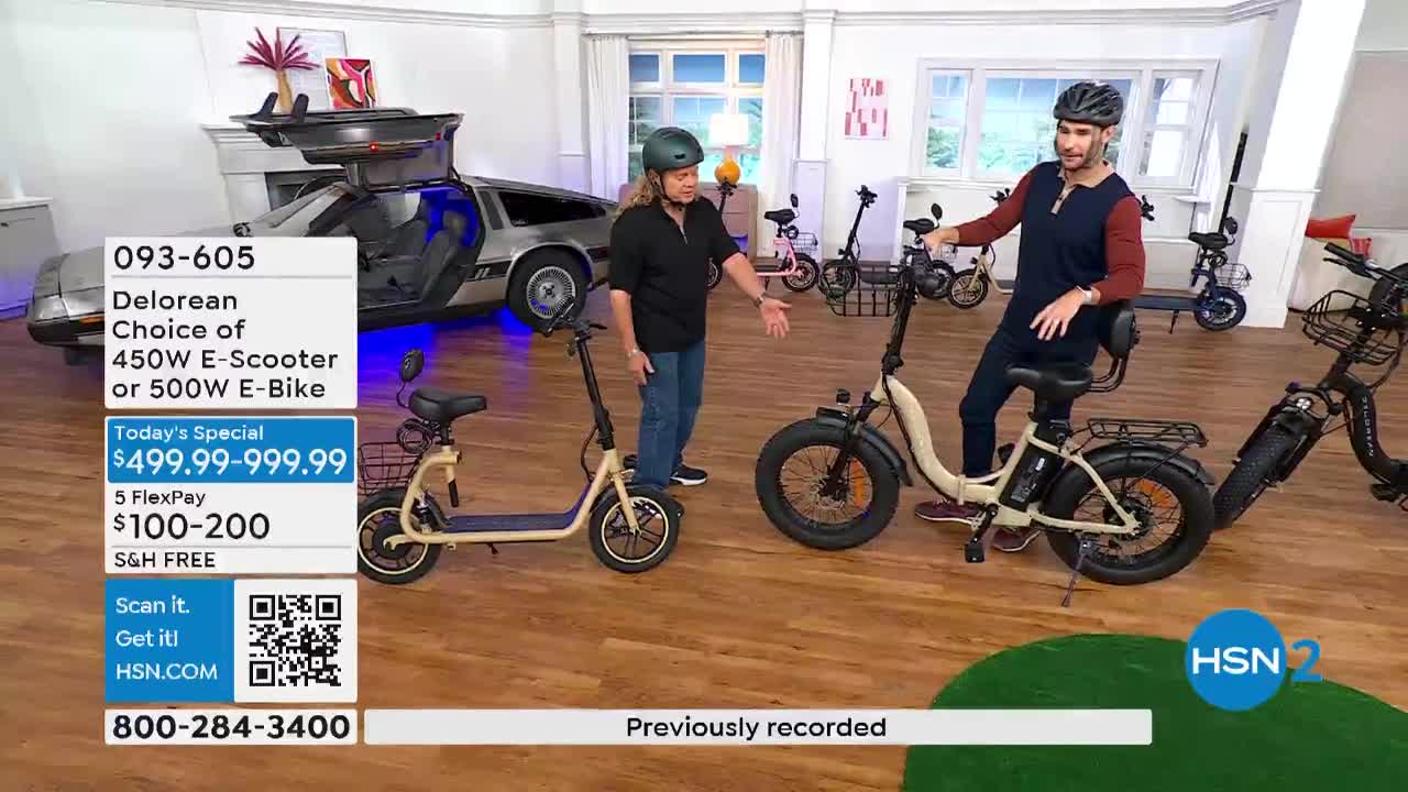Two men, both wearing helmets, stand beside different models of electric bikes on a set for HSN2. One man gestures at the bikes, while the other sits on one, and a DeLorean sits in the background.
