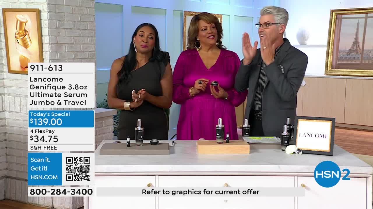 Three people stand behind a table, gesturing toward the products on display. The man in the gray suit claps his hands, while the women hold small bottles, all of them smiling.
