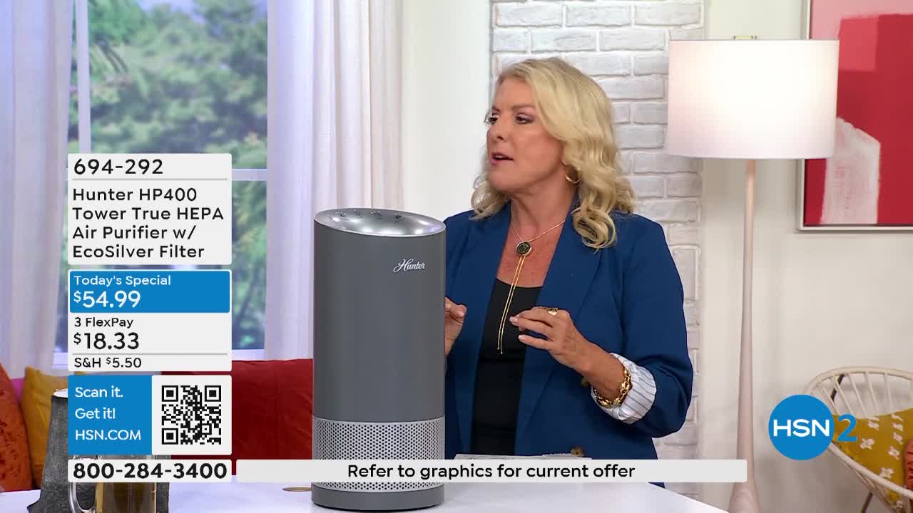 A woman in a blue blazer gestures toward a tall, gray air purifier. The HSN2 logo is visible in the corner, and the screen displays the product's price.
