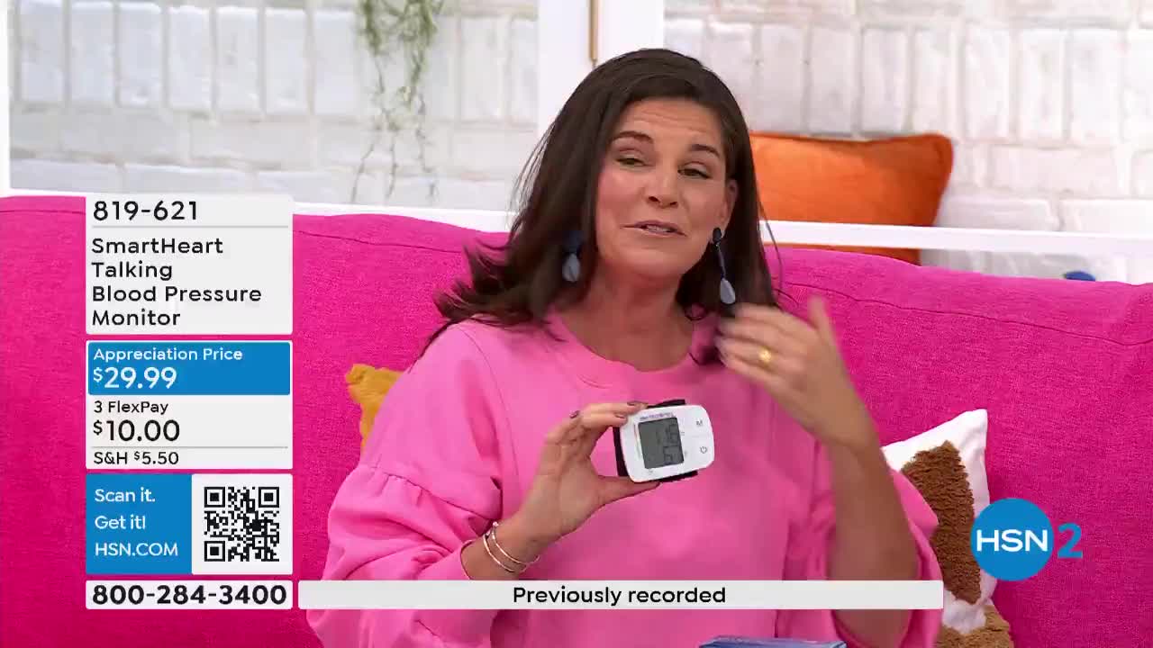 A woman in a pink sweater holds up a small, white blood pressure monitor, gesturing with her other hand. The HSN2 logo is visible in the corner, and the screen displays product information.
