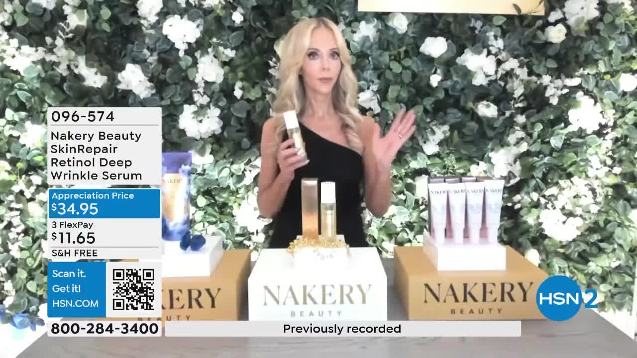 A woman in a black dress gestures towards a bottle of serum, holding one in her hand. The set, on HSN2, is decorated with white flowers and displays the product's name, Nakery Beauty.
