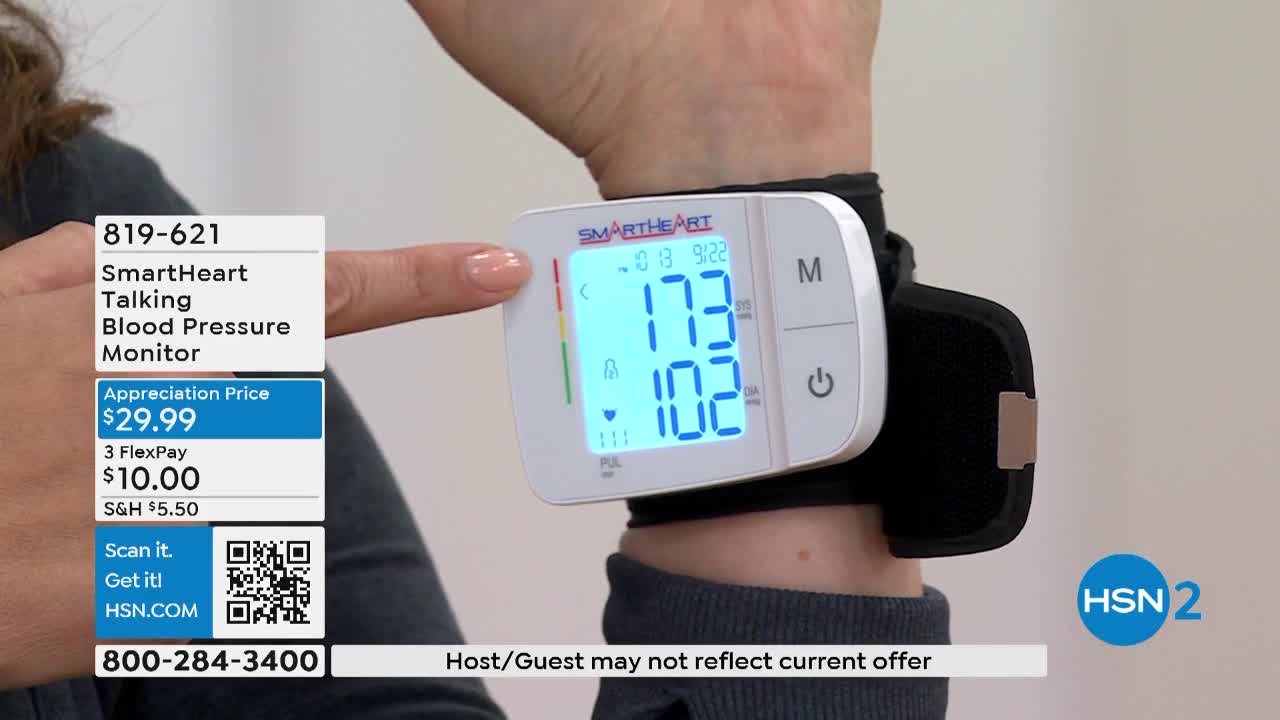 A person's finger points to a color-coded bar on a SmartHeart blood pressure monitor strapped to their wrist. The monitor displays a reading of 173/102 on the HSN2 broadcast.
