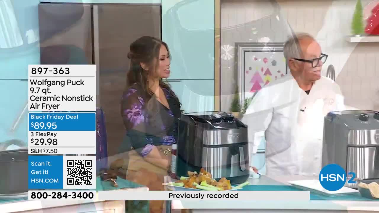 A woman in a floral top smiles as a chef in a white coat gestures toward an air fryer. On a counter, a plate of chicken wings and celery sits next to the appliance.
