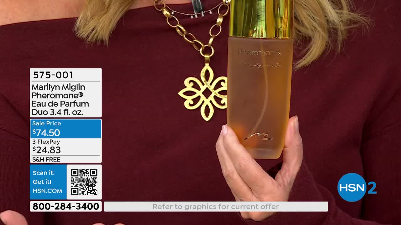 A woman holds a perfume bottle, its amber liquid visible through the frosted glass.  The HSN2 logo is in the corner, and a price tag is displayed on the screen.
