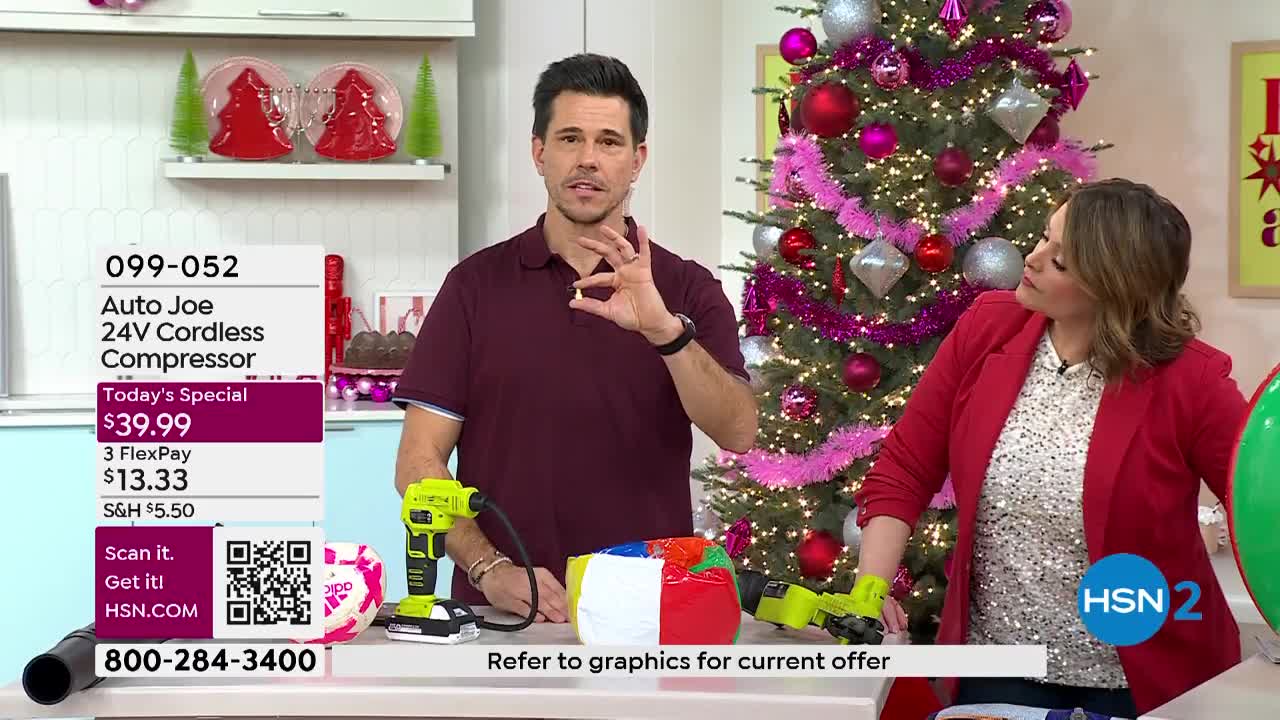 A man in a maroon shirt holds up a small object, gesturing with his hand. Beside him, a woman in a red blazer looks on, while a Christmas tree sparkles in the background.
