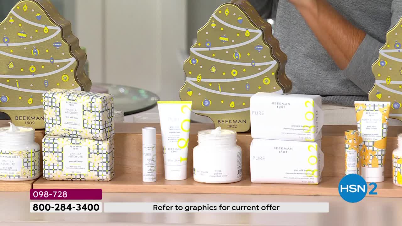 A collection of Beekman 1802 products, including soaps and creams, is arranged on a wooden shelf. Behind them, festive tree-shaped boxes add a touch of holiday flair to the HSN2 presentation.
