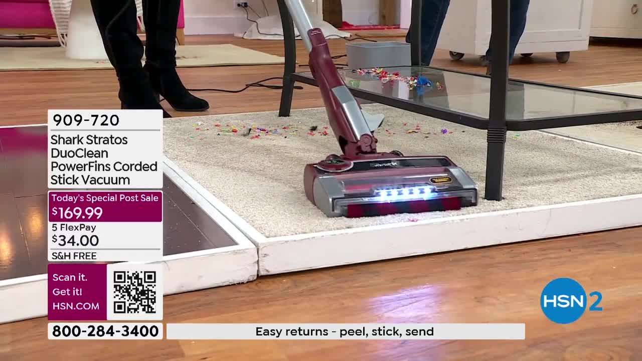 A maroon and silver vacuum cleaner is moving across a carpeted platform, illuminated by its bright headlights. Scattered debris is being sucked up, and the HSN2 logo is visible in the corner.
