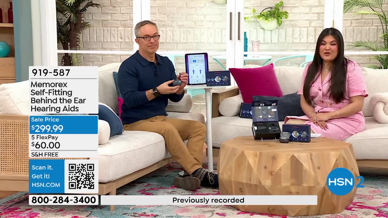 A man holds up a small, dark gray device, gesturing towards a tablet screen displaying hearing aid settings. Beside him, a woman in a pink dress smiles as she sits on a couch, with several product boxes arranged on a wooden table.