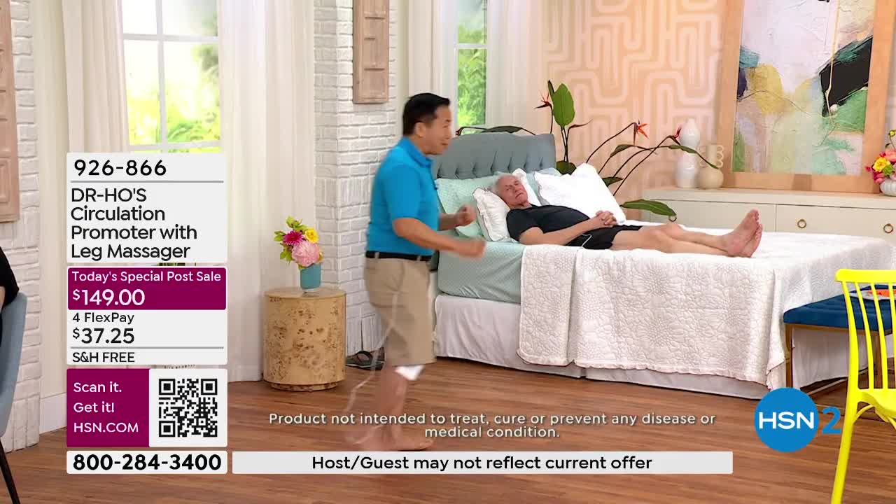 A man in a blue shirt gestures as he stands next to a bed where another man lies. A device is attached to the standing man's leg.