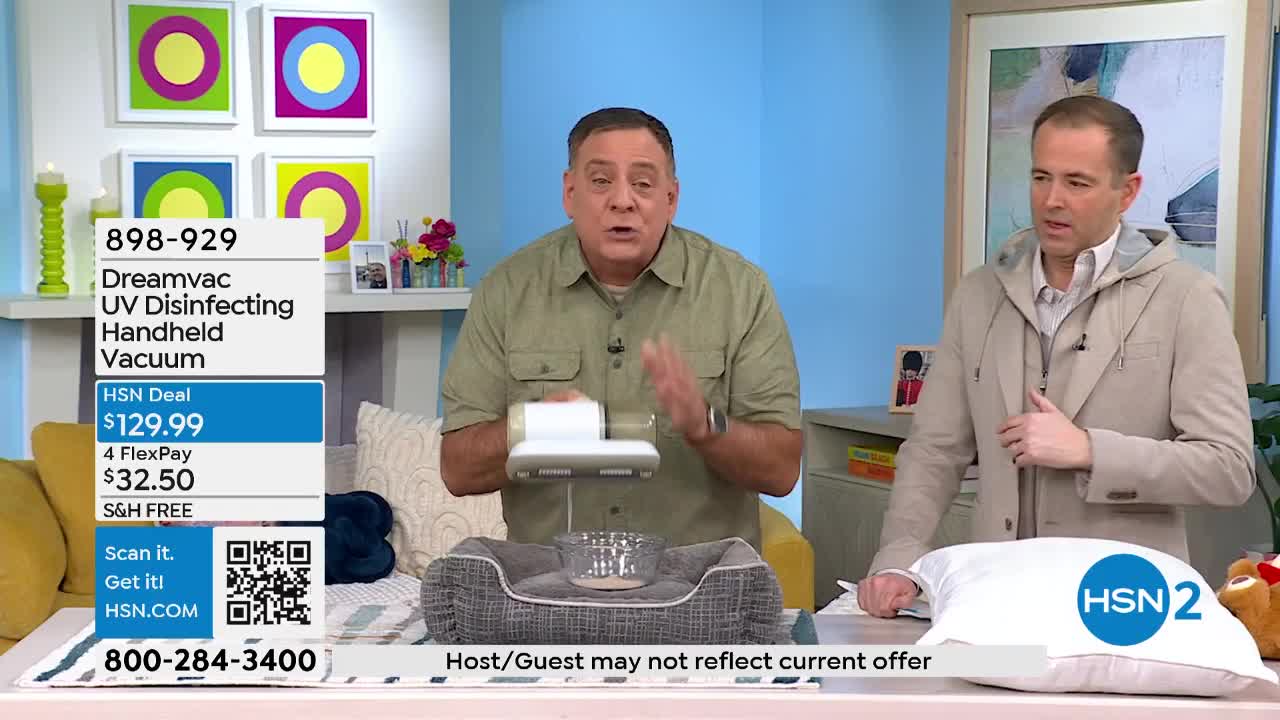 A man in a green shirt demonstrates a handheld vacuum over a pet bed. Another man in a tan jacket stands beside him, looking on.