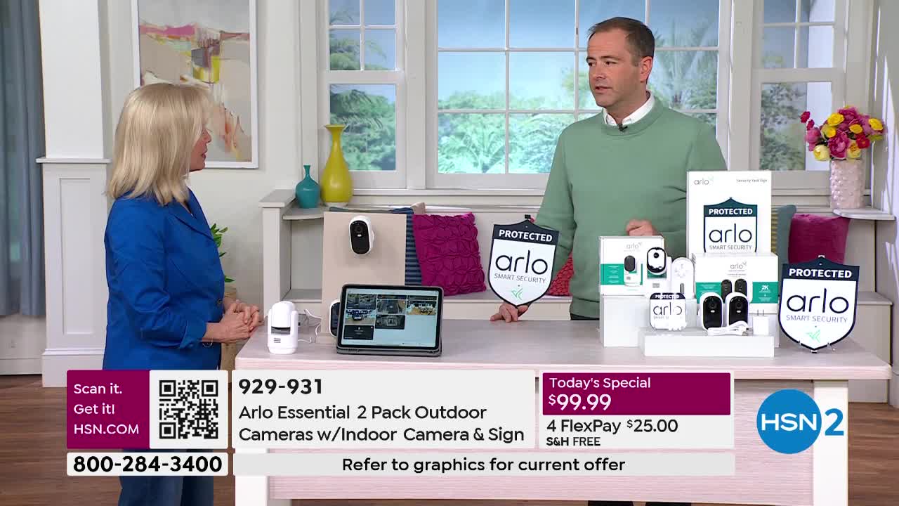 A woman in a blue jacket stands to the left, facing a man in a green sweater. He gestures towards a display of Arlo security cameras on a table.