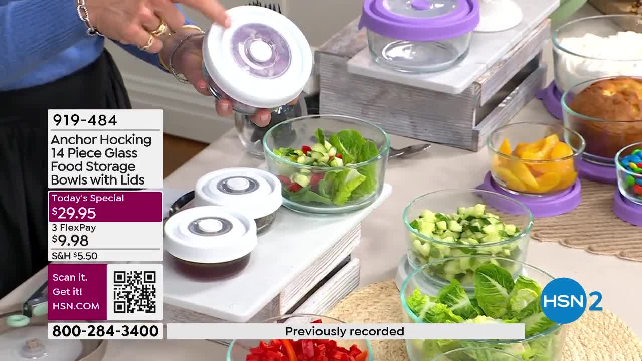 A hand holds up a clear glass bowl with a white lid, showing off its vacuum seal. Several other bowls filled with fresh ingredients like chopped cucumbers and salad greens are arranged on a table.
