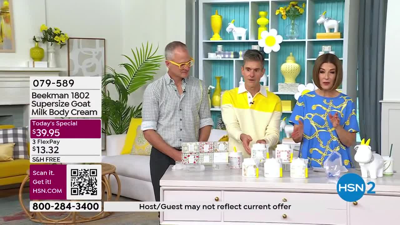 A woman in a blue dress gestures enthusiastically while a man in a yellow shirt stands beside her, both presenting jars of Beekman 1802 body cream on a table. The HSN2 logo is visible in the lower right corner.