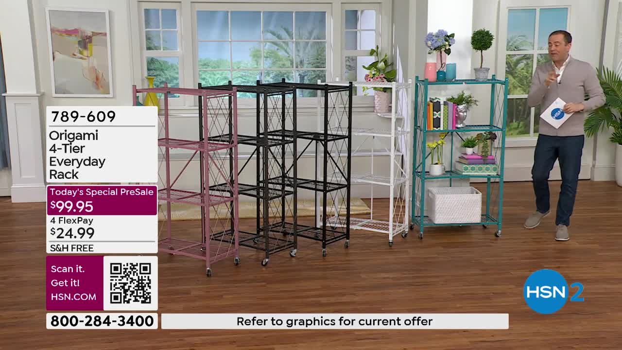 A man in a grey sweater and blue jeans gestures as he stands next to a teal shelving unit. Four foldable racks, in pink, black, white, and teal, are displayed on a wooden floor.