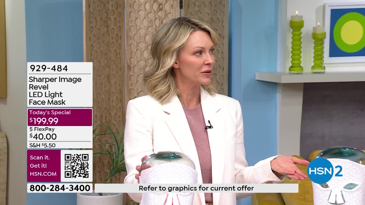 A woman in a white blazer gestures towards a clear dome with a white, patterned base. A graphic overlay displays "Sharper Image Revel LED Light Face Mask" and pricing information.