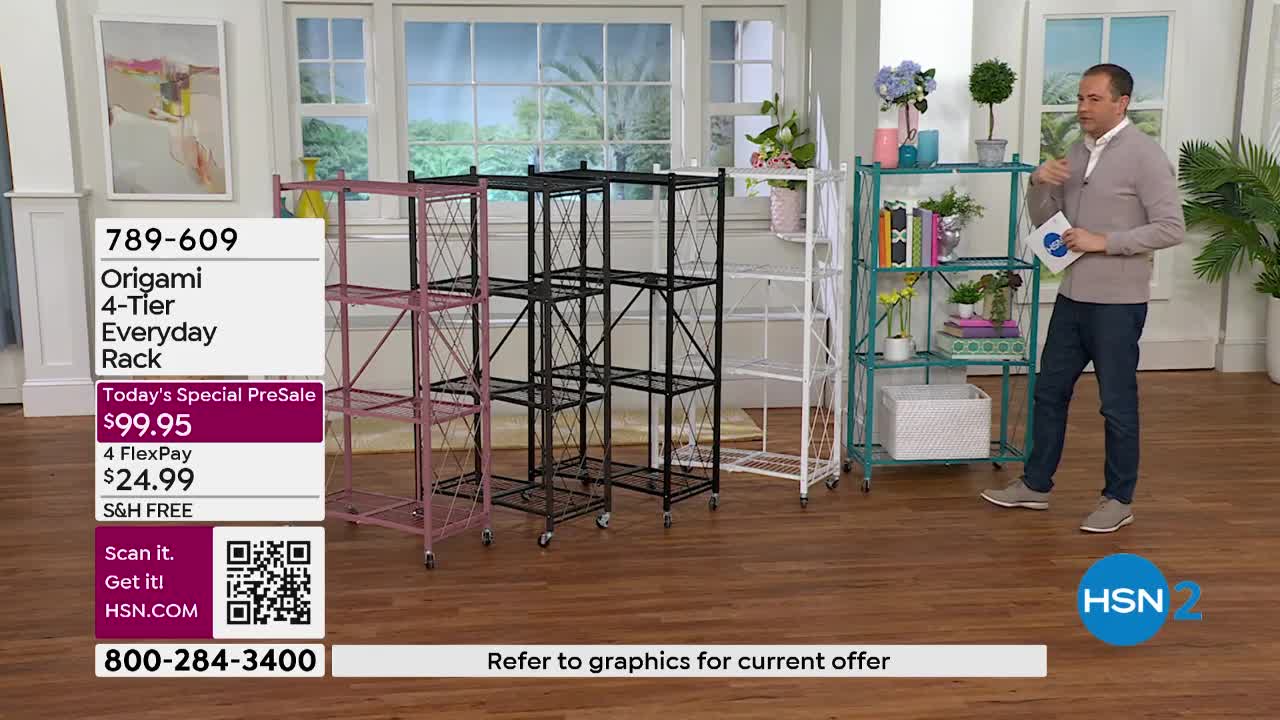 A man stands by a teal shelving unit, gesturing with a white card. Several Origami racks, in pink, black, and white, are lined up on the floor.