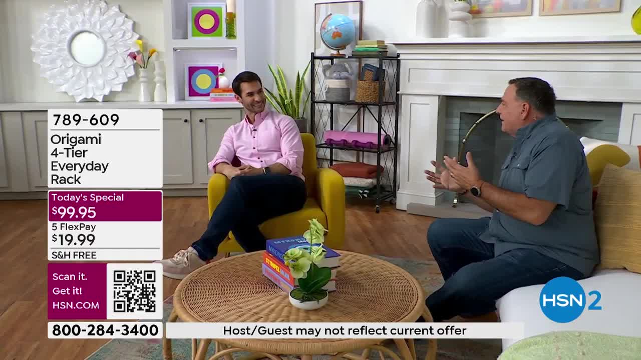 Two men are seated in a living room set, discussing an Origami 4-Tier Everyday Rack being presented on HSN2. One man, wearing a pink shirt, gestures animatedly while the other, in a blue shirt, listens intently.