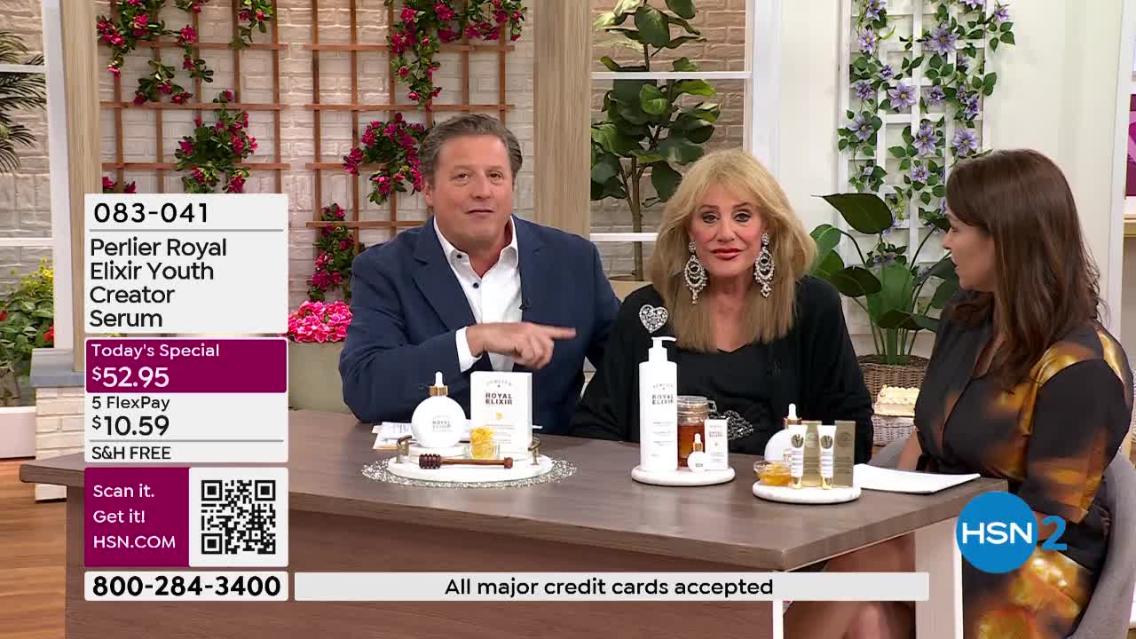 A man in a blue suit gestures towards a display of Perlier Royal Elixir serum bottles. A woman with blonde hair and large earrings smiles beside him, while another woman in a patterned dress looks on.