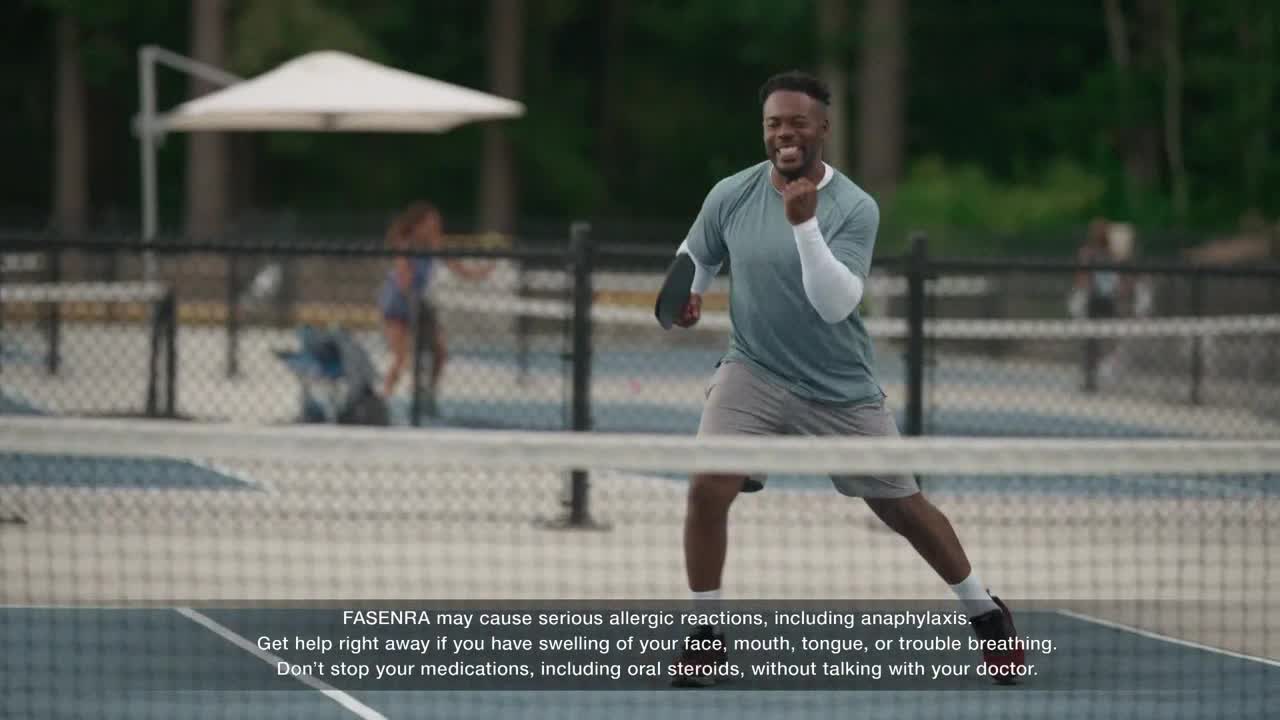 A man in athletic gear is mid-stride on a pickleball court, paddle in hand. He's smiling and appears to be enjoying himself, while a woman and a stroller are in the background.
