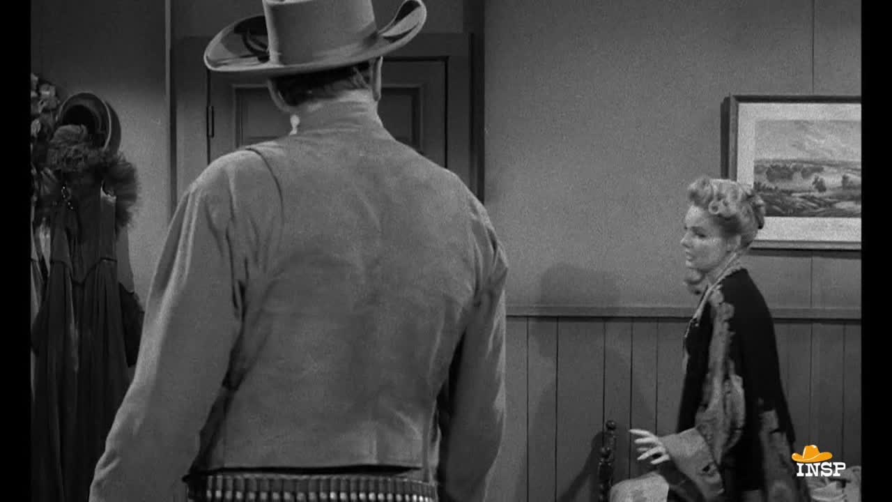 A man in a cowboy hat and vest stands with his back to us, facing a door. A woman in a patterned shawl gestures with her hand as she looks at him, her expression concerned.
