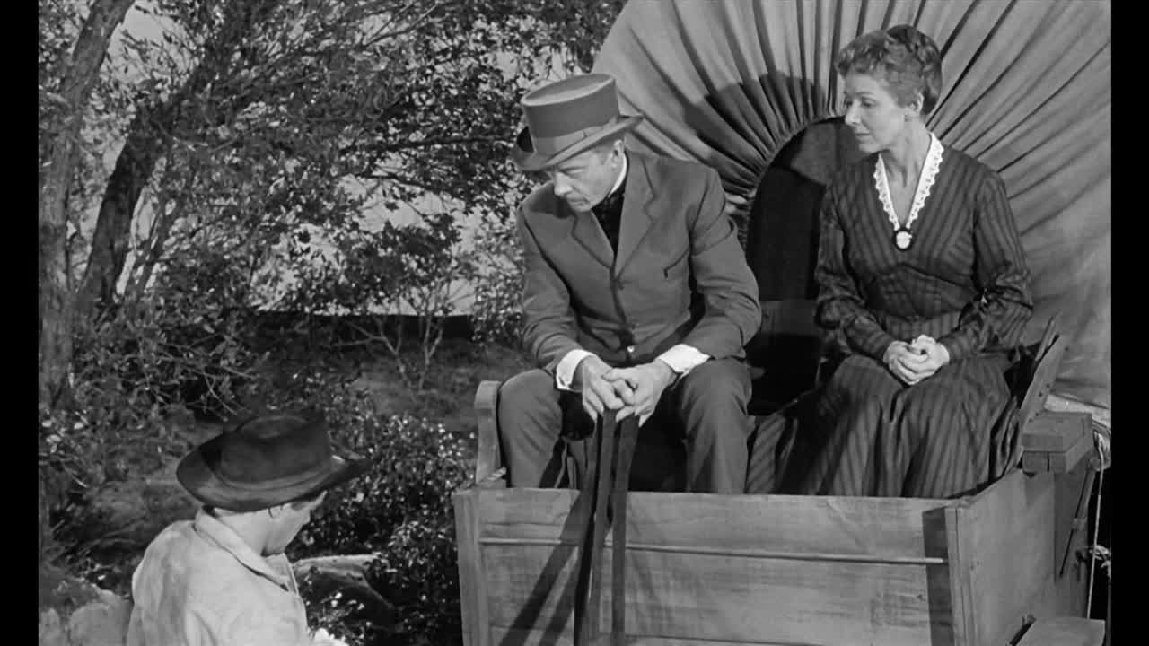 A man in a top hat sits with a woman in a wagon, both looking down. The man in the hat holds a cane, while another man in a cowboy hat stands nearby.
