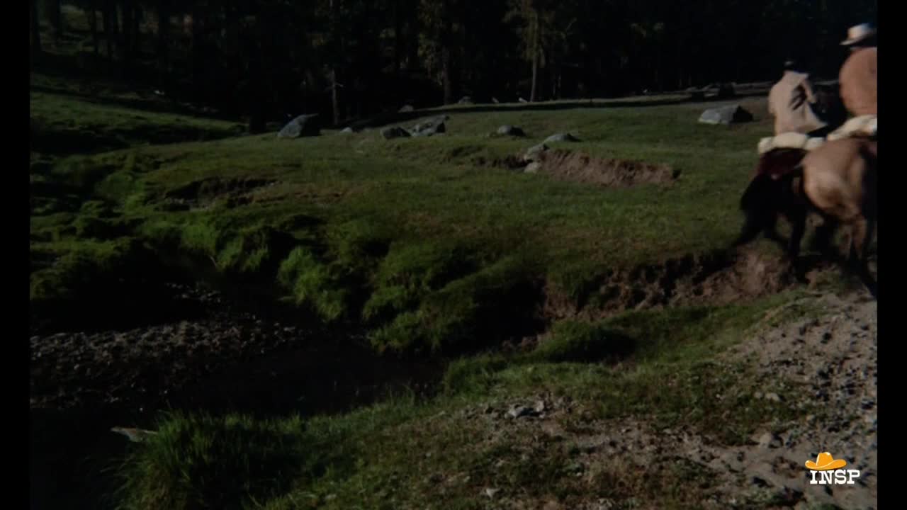 Two riders on horseback are approaching a small creek, the horses' dark coats contrasting with the green grass. The scene is set in a grassy field with a dark forest in the background, likely a scene from a Western on INSP.
