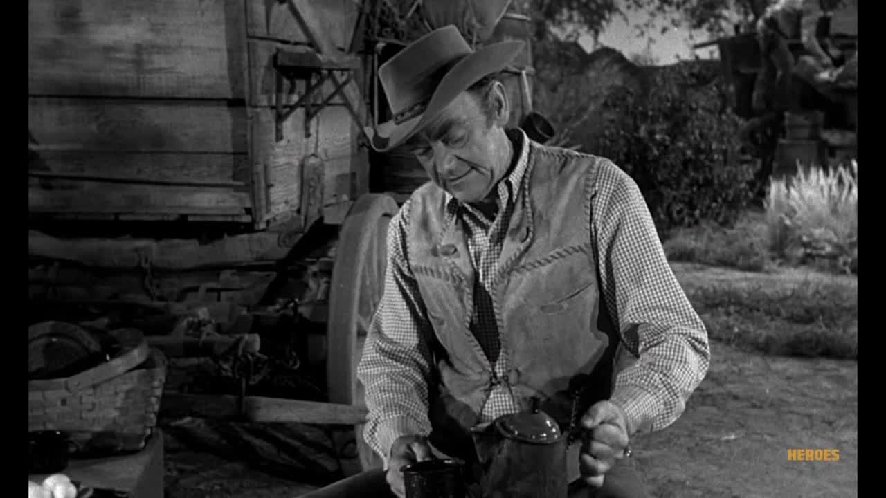 The man in the cowboy hat carefully pours from a dark metal pot. He's seated near a wooden structure, likely preparing a meal.
