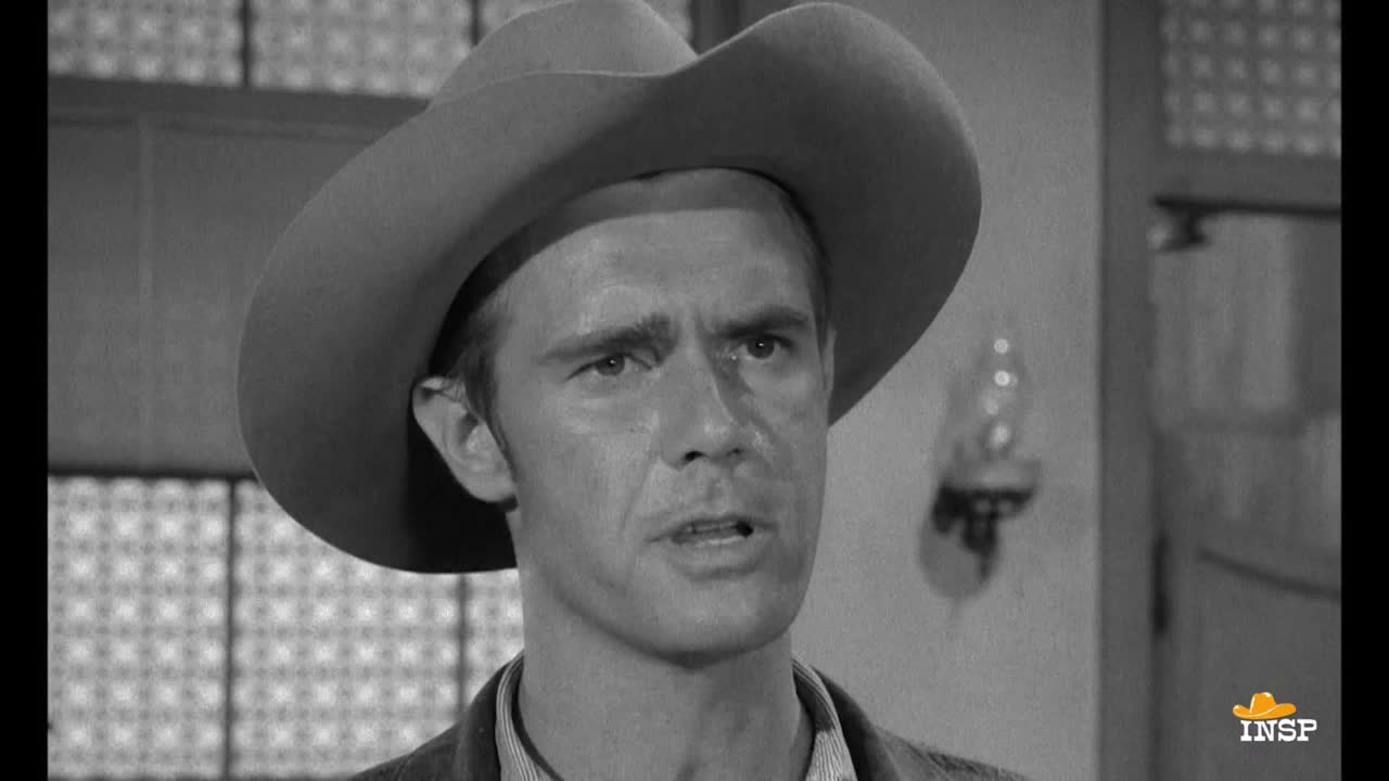 A man in a cowboy hat is speaking, his brow furrowed. The scene is a black and white Western, likely from INSP.

