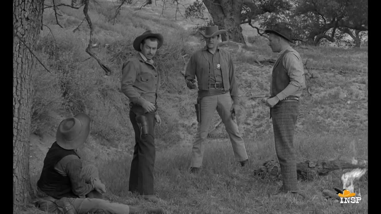 Four men in cowboy hats stand in a grassy area, one with a gun drawn. Another man sits on the ground, leaning against a tree.
