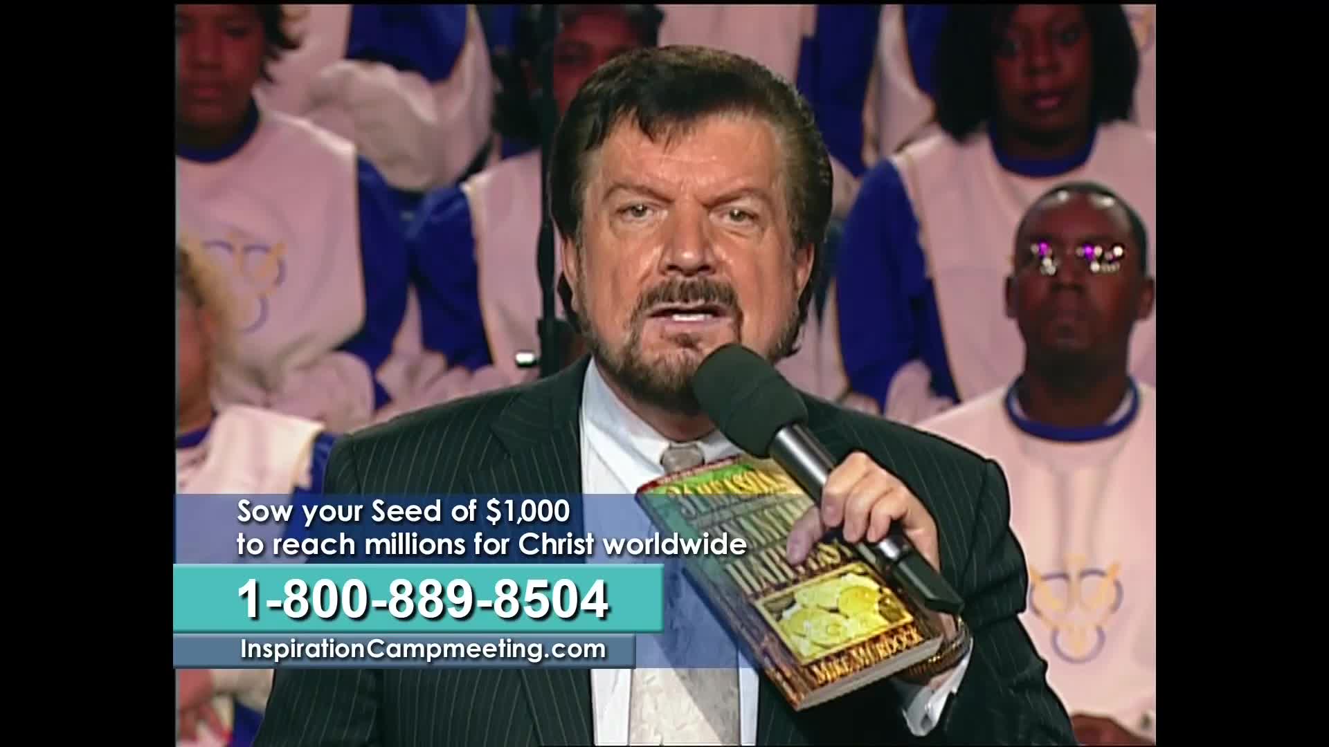 A man in a suit, holding a microphone and a book, addresses the camera on what looks like a television program. Behind him, a choir of people in white and blue robes are seated.
