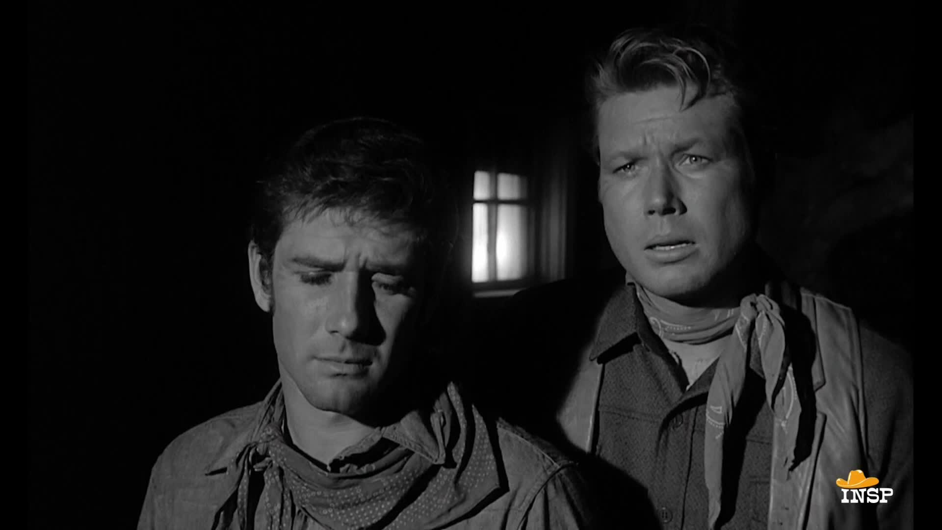 Two men stand close together, their faces lit dimly by a window behind them. The man on the right looks shocked, while the other appears to be lost in thought.
