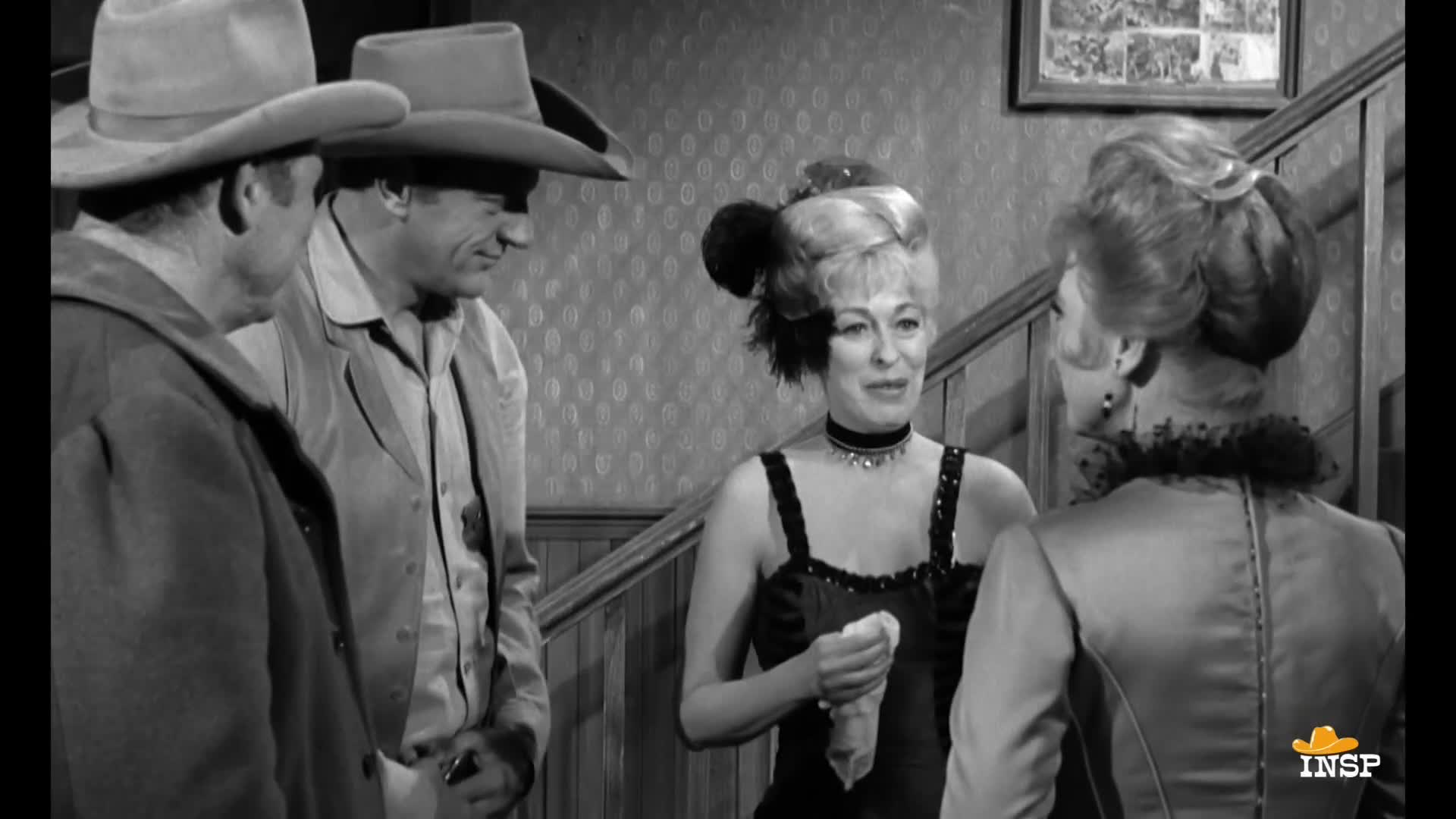 Two men in cowboy hats stand close, listening to a woman in a black dress holding a handkerchief. The woman on the right, in a high-collared dress, faces the group, and the scene is set in a classic Western saloon.
