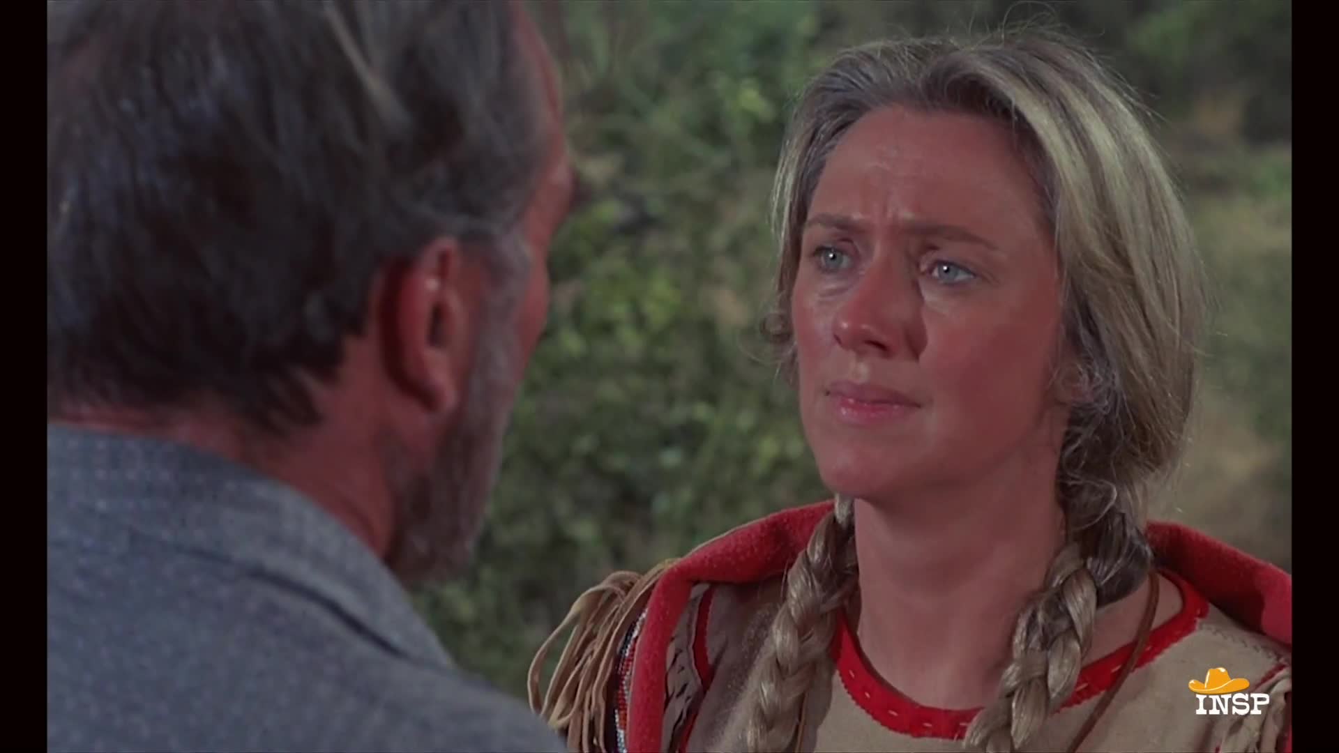 A woman with braided blonde hair is looking intently at a man, her face flushed. She's wearing a fringed, red-trimmed garment, and the man is turned away, his face partially obscured.
