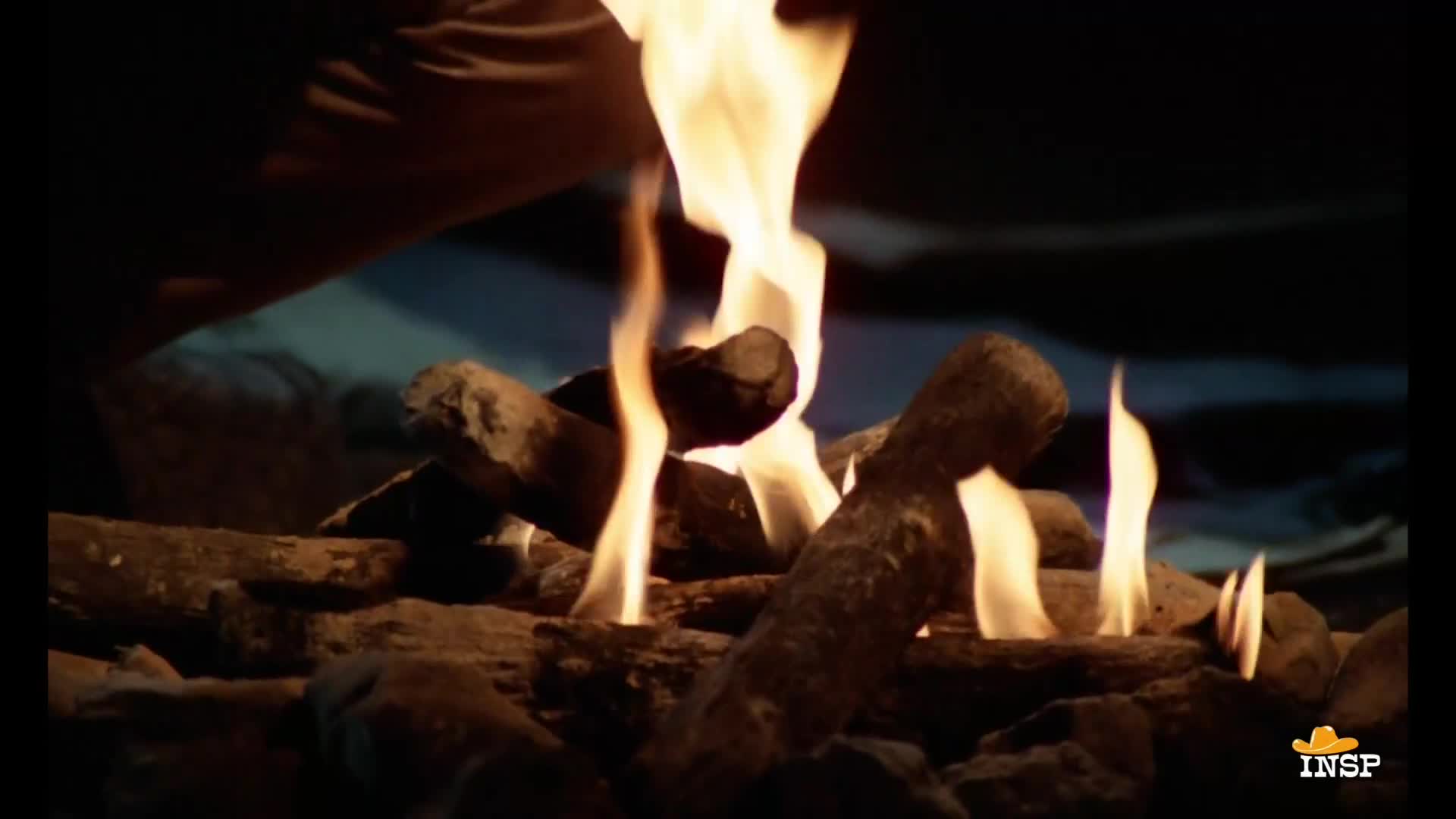 Flames dance upwards from a pile of logs, their orange and yellow tongues flickering against the dark backdrop. Someone is reaching down, perhaps to tend to the fire.
