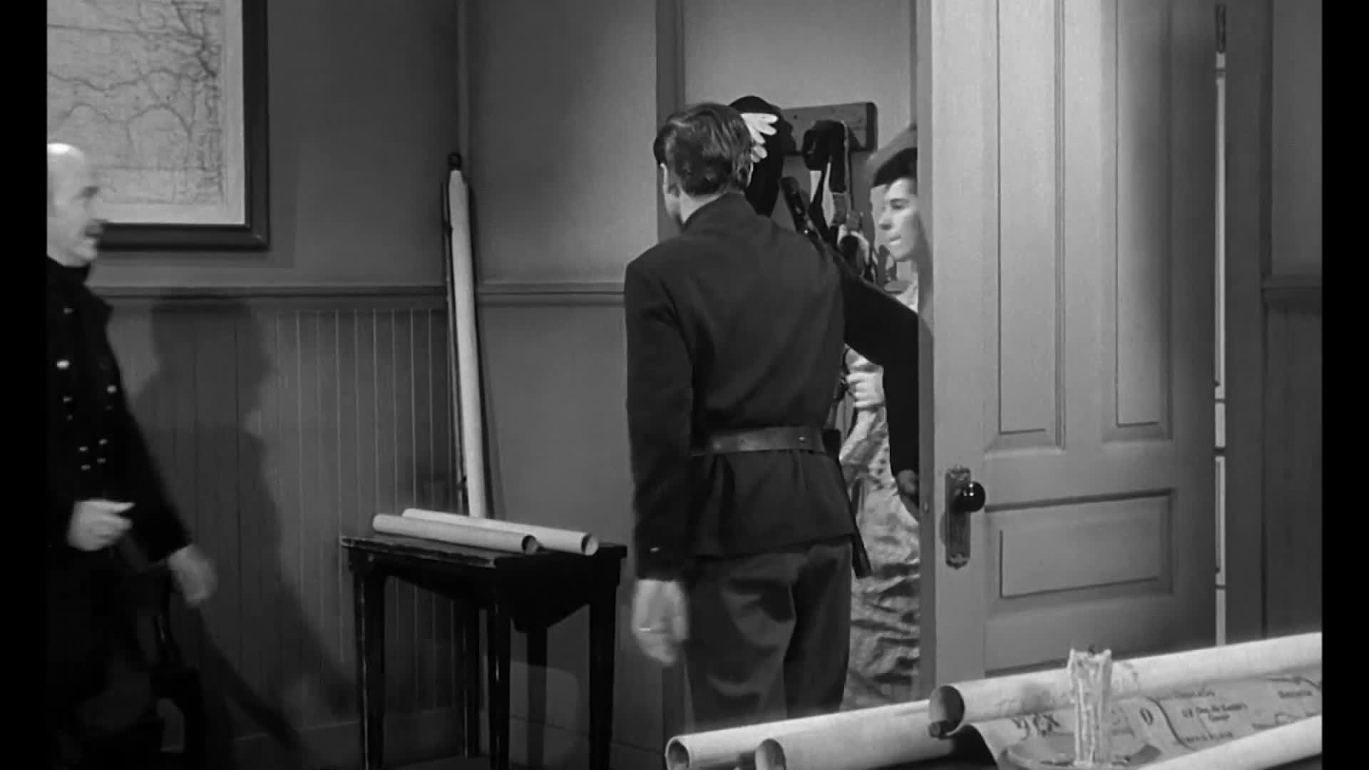 A man in a dark uniform walks toward a doorway, while a woman peeks out from behind it. Another man in uniform is exiting the room, his back to us.

