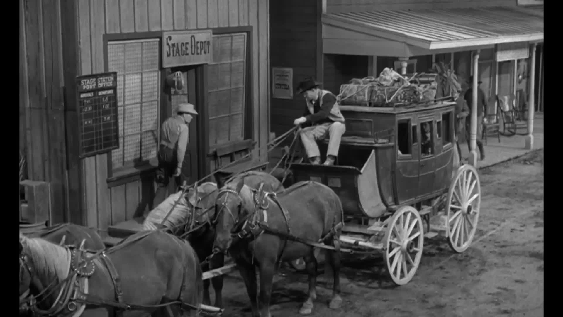 A stagecoach sits in front of a depot, ready to depart. The driver is perched on top, holding the reins, while a man near the door checks the schedule.
