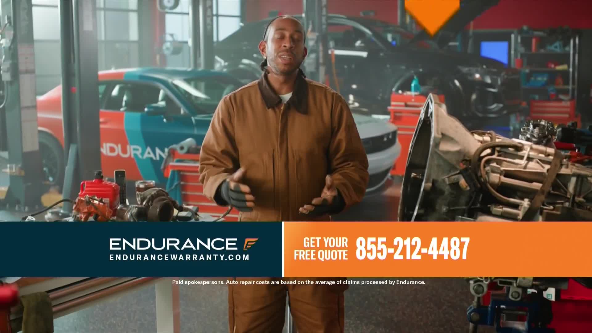 A man in a brown work jacket stands in a garage, gesturing with his hands. Behind him, a red and blue car is parked, and a large transmission sits on a nearby table.
