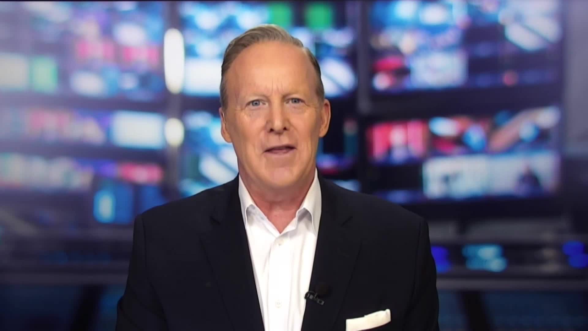 A man in a dark suit and white shirt is speaking, his expression open and direct. Behind him, a wall of screens displays various images, likely from a television studio.
