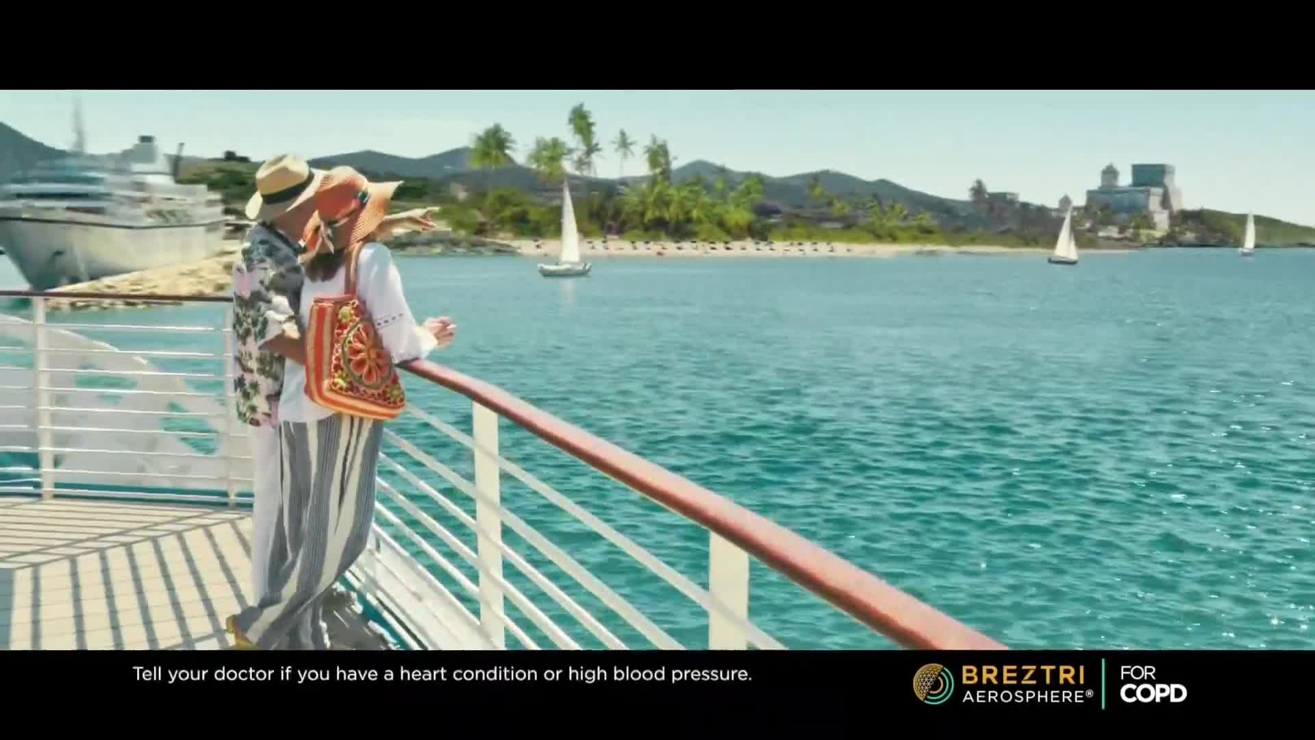A couple stands on a cruise ship deck, gazing out at the turquoise water and a distant island. The woman points towards the horizon, while sailboats drift peacefully across the bay.
