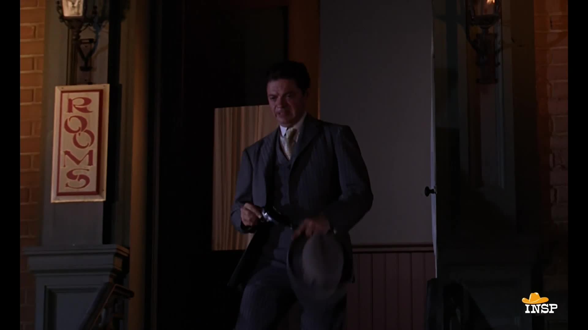 A man in a suit exits a doorway, holding a hat in one hand and a gun in the other. The "Rooms" sign hangs above the entrance.
