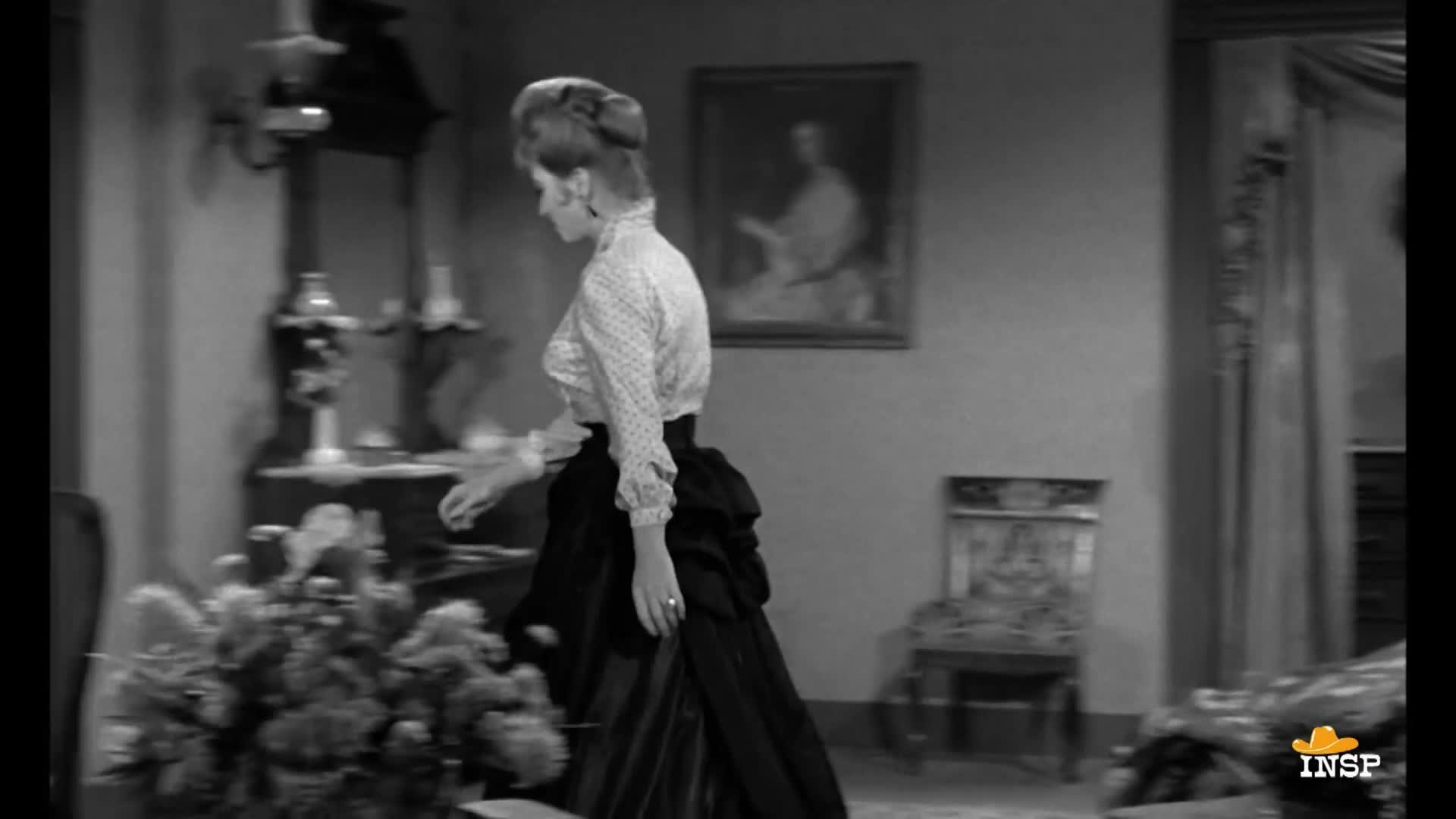 A woman in a patterned blouse and long skirt walks purposefully across the room. She glances down at a large floral arrangement as she passes.
