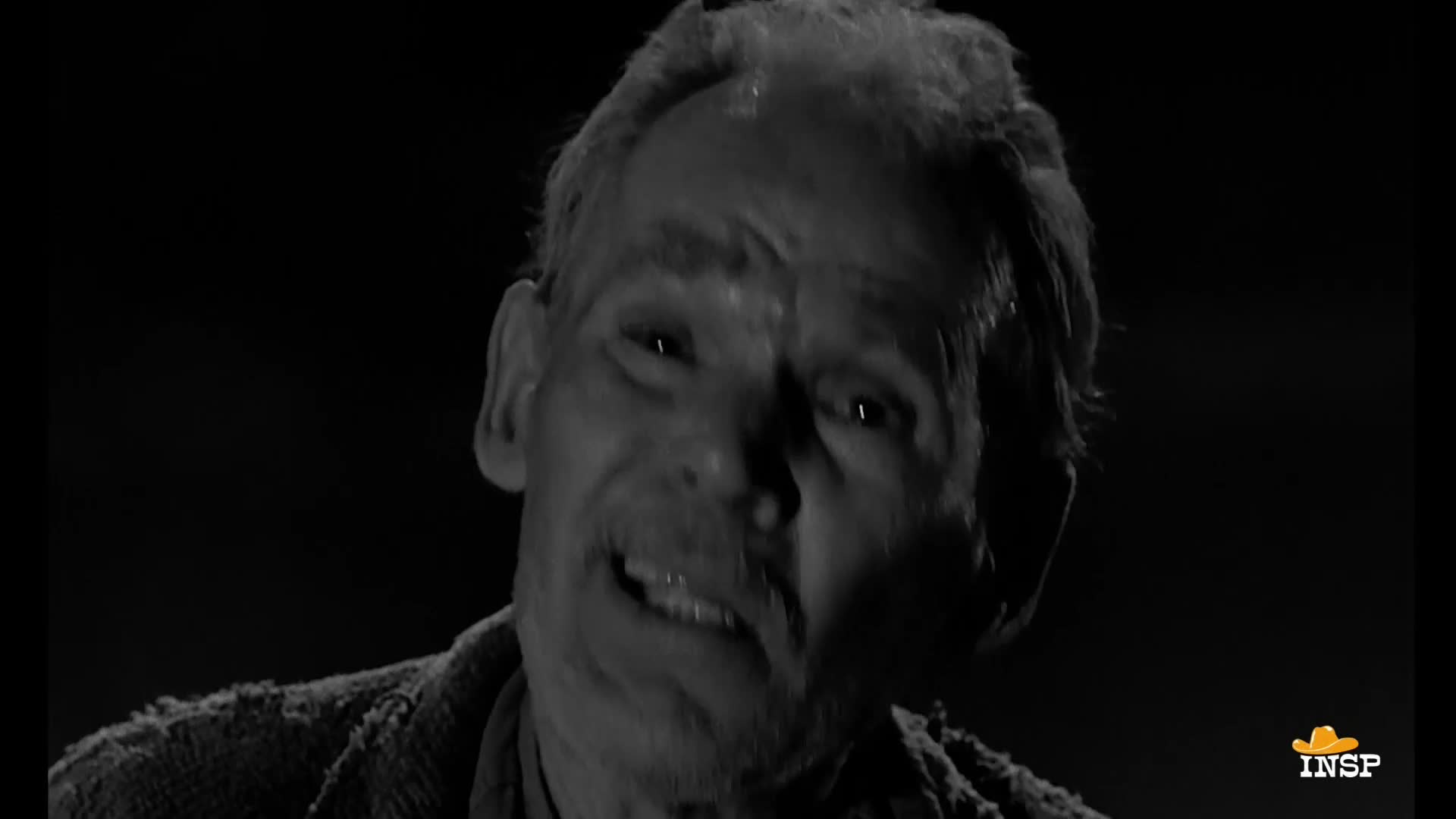 The man's face is lit, his eyes wide and mouth slightly open as if he's about to speak. He's wearing a textured, dark sweater against a black background, a scene from INSP.
