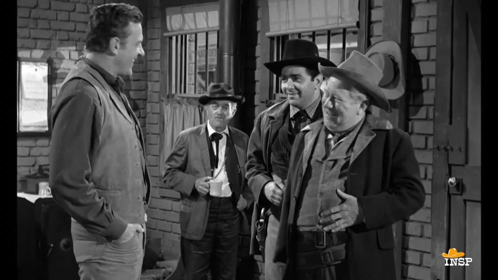 A man in a vest and jeans is chatting with a group of men, one of whom is holding a mug. The scene appears to be a Western, possibly a jailhouse, with the INSP logo in the corner.
