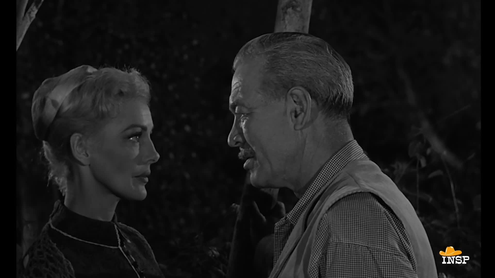 The woman, her blonde hair neatly styled, gazes intently at the man. He's turned towards her, his mouth slightly open as if he's about to speak.
