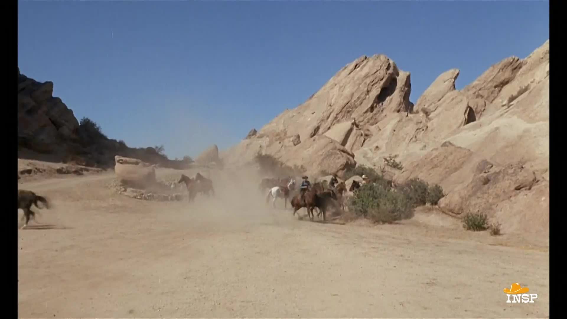 A herd of horses, ridden by cowboys, kicks up a cloud of dust as they race through a desert canyon. The riders are silhouetted against the sun, and the rocky landscape looms in the background.
