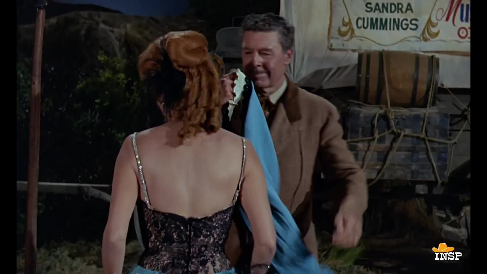 A man in a brown jacket holds out a blue cloth, offering it to a woman in a sparkling gown. Behind them, a sign advertises "Sandra Cummings."
