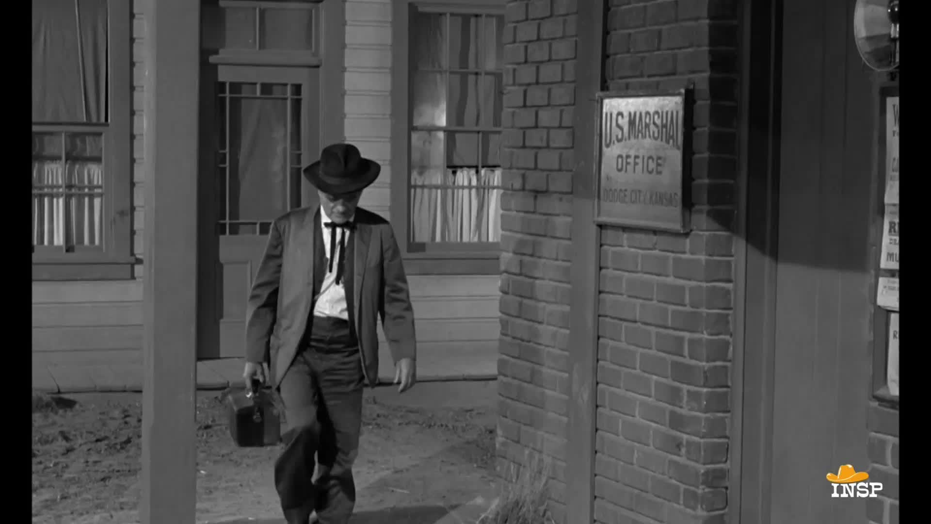 A man in a dark suit and hat walks away from the U.S. Marshal's office, a small case in his hand. He's headed down the street in Dodge City, Kansas.
