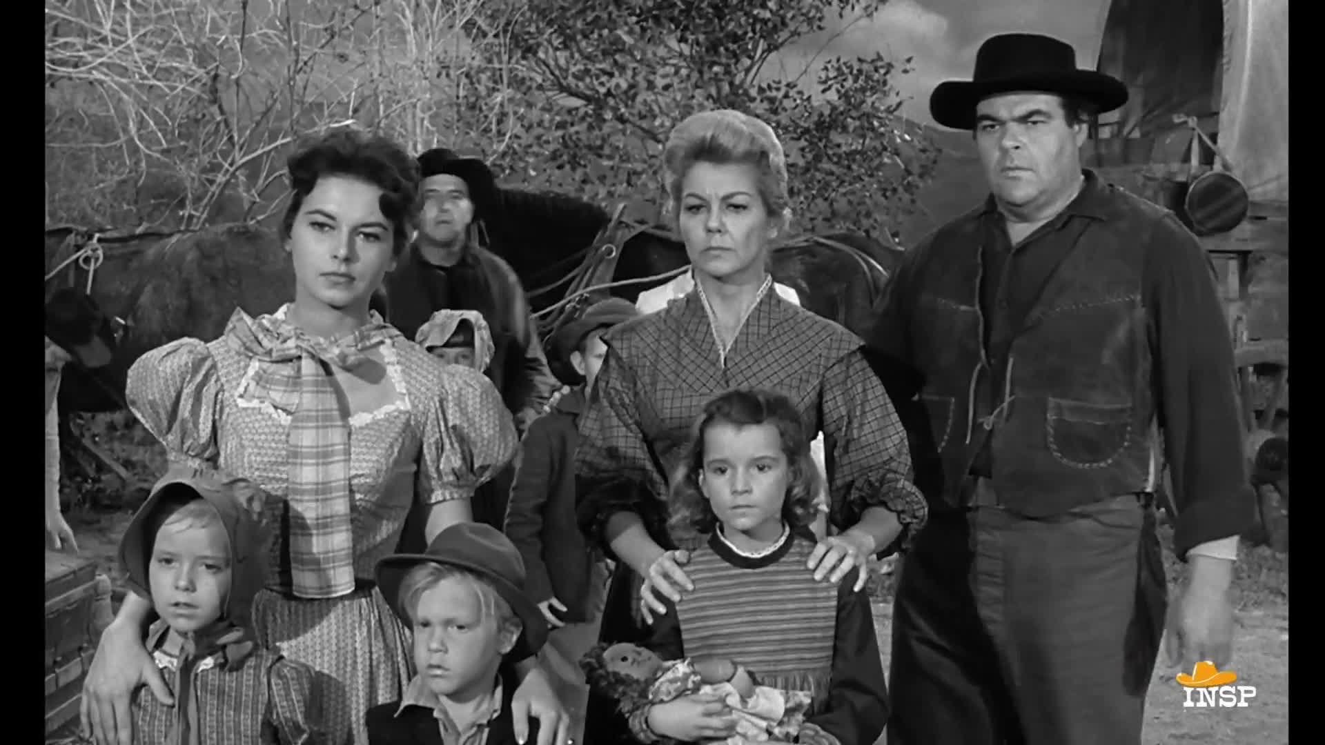 A group of people stands together, their faces serious, as if posing for a portrait. Behind them, a wagon and horses are visible, suggesting a journey or a departure.
