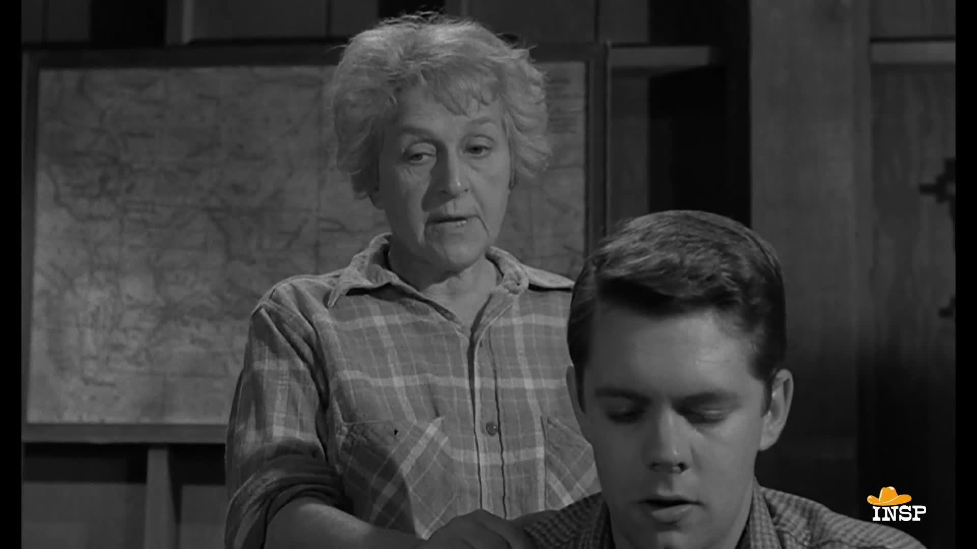 A woman in a plaid shirt leans over a young man, speaking. Behind them, a map hangs on the wall.
